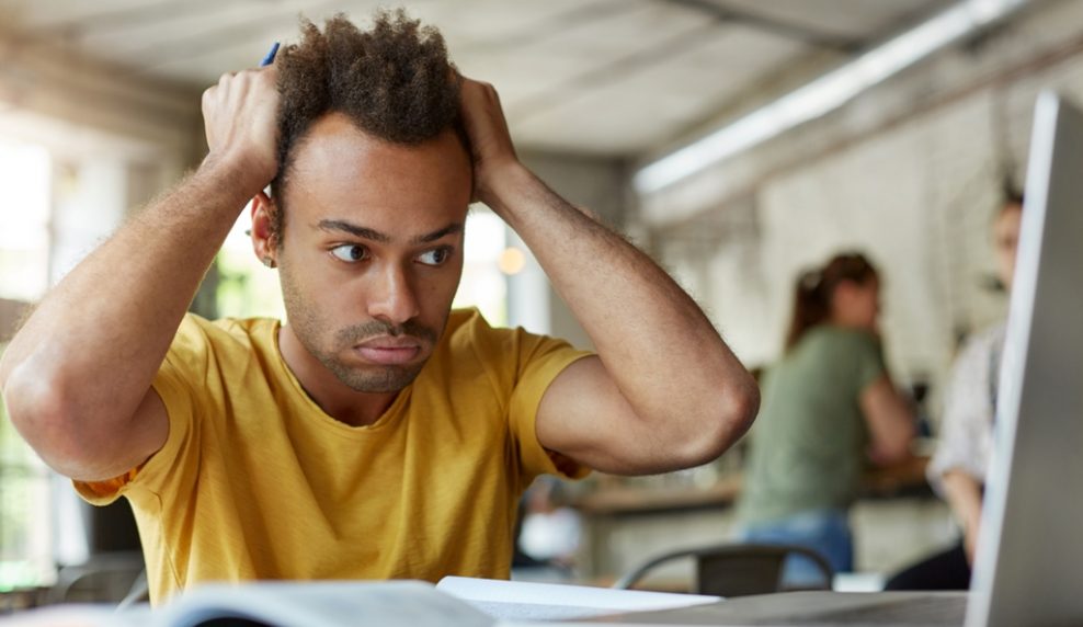 Stressed young African American student of school of economics feeling frustrated while working on diploma project, sitting at coworking space in front of open laptop computer, holding head with hands
